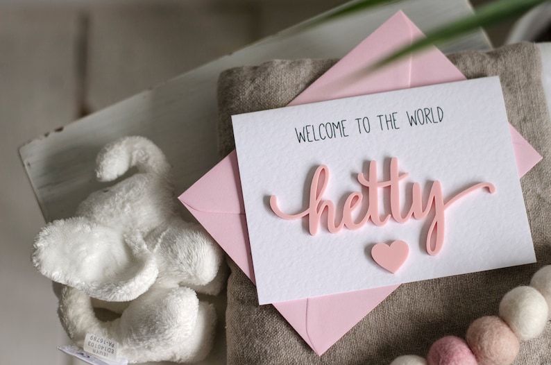 May include: A pink card with the text "Welcome to the world" and the name "Hetty" in pink raised letters. A small pink heart is below the name.