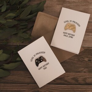 May include: Two white greeting cards with a black and white illustration of a video game controller. The text on the cards reads "Level 10 Unlocked! Happy Birthday Theo" and "Level 33 Unlocked! Happy Birthday Uncle Jimbo".