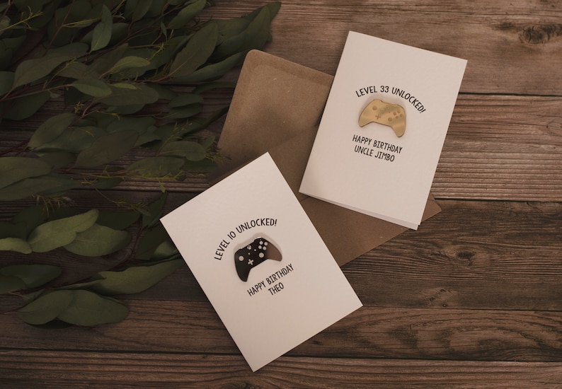 May include: Two white greeting cards with a black and white illustration of a video game controller. The text on the cards reads "Level 10 Unlocked! Happy Birthday Theo" and "Level 33 Unlocked! Happy Birthday Uncle Jimbo".