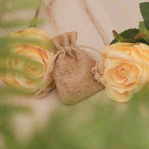 May include: Two yellow roses and a small burlap pouch with a drawstring closure. The roses are in focus, while the pouch is slightly out of focus. The background is a light brown surface.