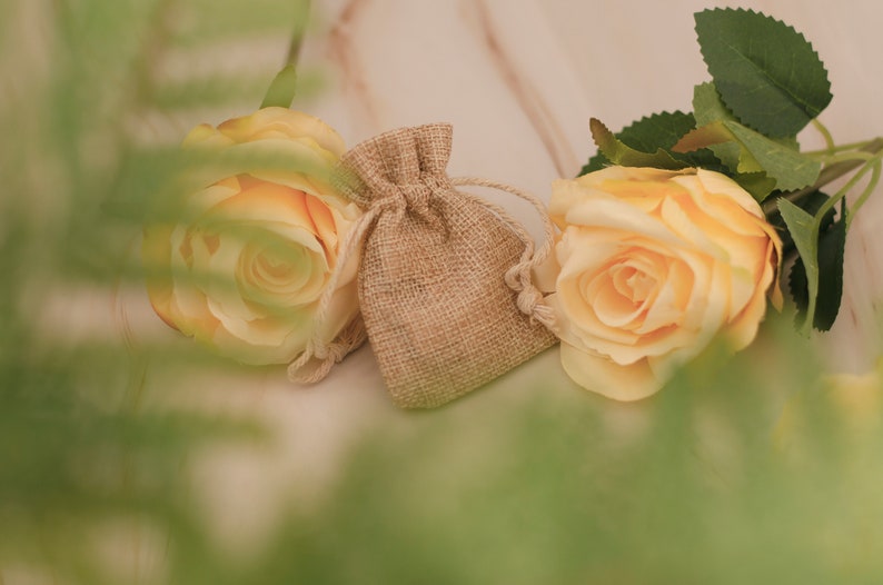 May include: Two yellow roses and a small burlap pouch with a drawstring closure. The roses are in focus, while the pouch is slightly out of focus. The background is a light brown surface.