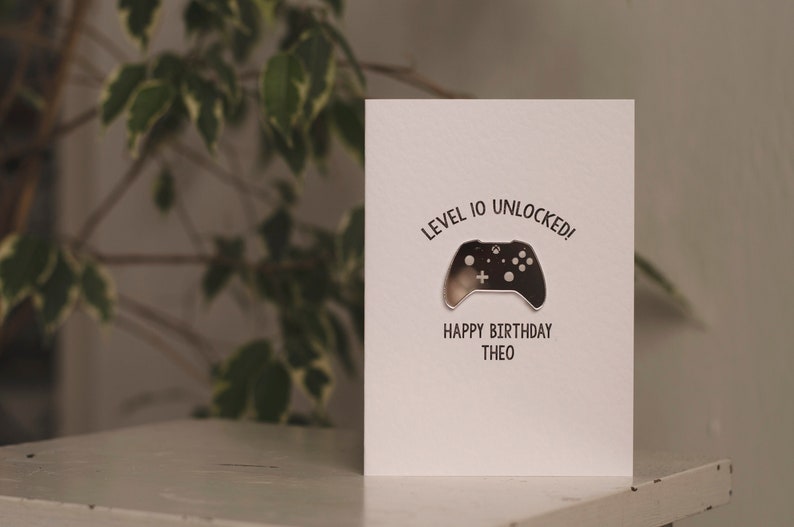 May include: A white card with the text "Level 10 Unlocked!" and a silver video game controller graphic. The text "Happy Birthday Theo" is below the graphic.
