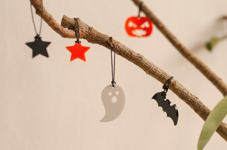 May include: A branch with five Halloween-themed decorations hanging from it. The decorations are a black star, a red star, a white ghost, a black bat, and an orange pumpkin.