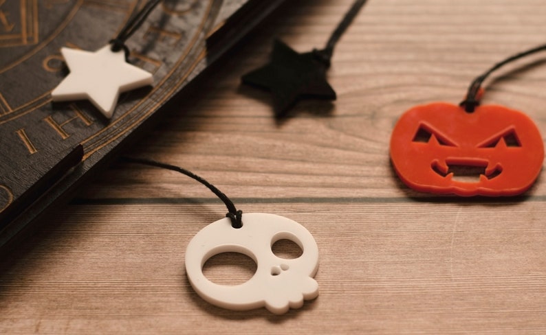 May include: Four Halloween-themed charms on black cords. The charms are a white star, a black star, a white skull, and an orange pumpkin with a jack-o'-lantern face.