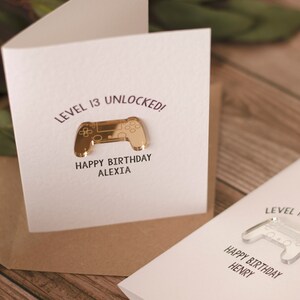 May include: Two white greeting cards with a gold and clear plastic video game controller on each. The text on the cards reads "Level 13 Unlocked! Happy Birthday Alexia" and "Level 17 Unlocked! Happy Birthday Henry".