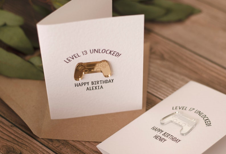 May include: Two white greeting cards with a gold and clear plastic video game controller on each. The text on the cards reads "Level 13 Unlocked! Happy Birthday Alexia" and "Level 17 Unlocked! Happy Birthday Henry".