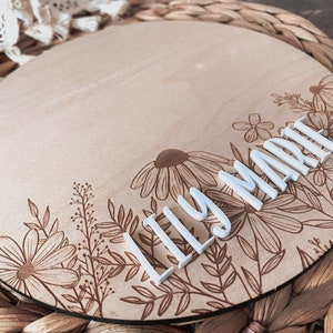 May include: A round wooden sign with a floral design and the name "Lily Marie" in white letters.