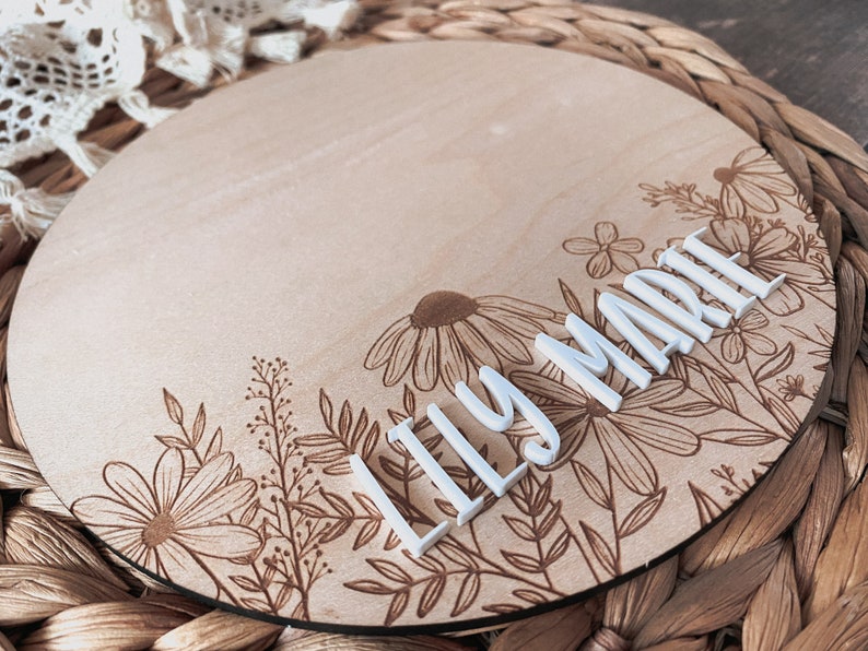 May include: A round wooden sign with a floral design and the name "Lily Marie" in white letters.