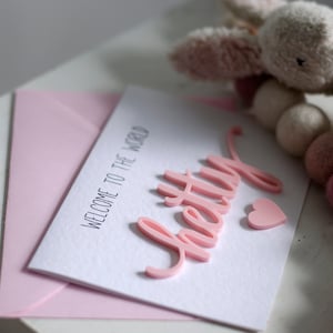 May include: A pink card with the text "Welcome to the world" and the word "pretty" in pink raised letters. A pink heart is next to the word "pretty".