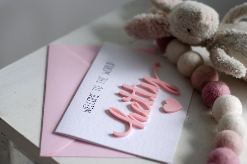 May include: A pink card with the text "Welcome to the world" and the word "pretty" in pink raised letters. A pink heart is next to the word "pretty".