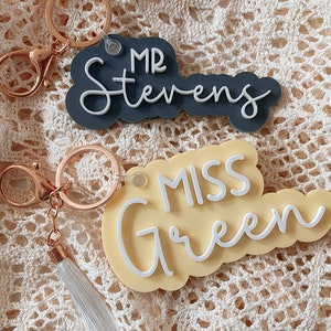 May include: Two keychains with personalised names. The first keychain is grey with white text that reads "MR Stevens". The second keychain is yellow with white text that reads "MISS Green". Both keychains have a gold-coloured chain and a tassel.