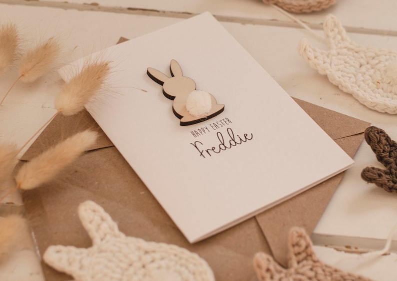 K&ouml;nnte beinhalten: Eine wei&szlig;e Karte mit einem ausgeschnittenen Holzhasen mit einem wei&szlig;en Bommel-Schwanz. Die Karte sagt "Frohe Ostern Freddie".
