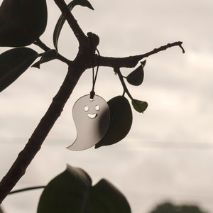 May include: A silver ghost-shaped charm with a smiling face hangs from a branch of a leafy plant.