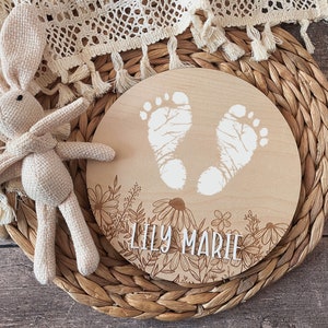 May include: A round wooden plaque with a floral design and two white baby footprints. The plaque is engraved with the name "Lily Marie".