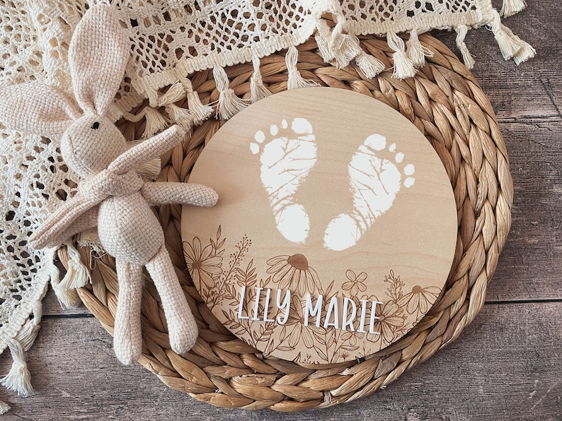 May include: A round wooden plaque with a floral design and two white baby footprints. The plaque is engraved with the name "Lily Marie".