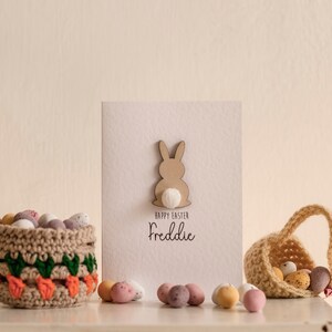 K&ouml;nnte beinhalten: Eine wei&szlig;e Karte mit einem braunen Hasen-Ausschnitt und dem Text "Frohe Ostern Freddie". Zwei geh&auml;kelte K&ouml;rbe gef&uuml;llt mit Schokoladeneiern stehen auf einer wei&szlig;en Oberfl&auml;che.