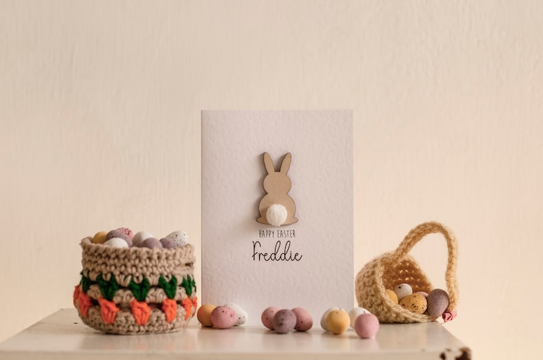 K&ouml;nnte beinhalten: Eine wei&szlig;e Karte mit einem braunen Hasen-Ausschnitt und dem Text "Frohe Ostern Freddie". Zwei geh&auml;kelte K&ouml;rbe gef&uuml;llt mit Schokoladeneiern stehen auf einer wei&szlig;en Oberfl&auml;che.