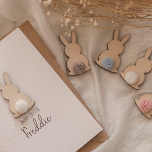 K&ouml;nnte beinhalten: Eine wei&szlig;e Karte mit dem Text "Happy Easter Freddie" und einer ausgeschnittenen Holzfigur eines Hasen mit einem wei&szlig;en Bommel-Schwanz. Vier weitere ausgeschnittene Holzfiguren von Hasen mit unterschiedlichfarbigen Bommel-Schw&auml;nzen sind auf einer wei&szlig;en Oberfl&auml;che verstreut.