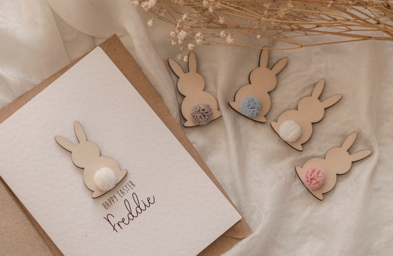 K&ouml;nnte beinhalten: Eine wei&szlig;e Karte mit dem Text "Happy Easter Freddie" und einer ausgeschnittenen Holzfigur eines Hasen mit einem wei&szlig;en Bommel-Schwanz. Vier weitere ausgeschnittene Holzfiguren von Hasen mit unterschiedlichfarbigen Bommel-Schw&auml;nzen sind auf einer wei&szlig;en Oberfl&auml;che verstreut.