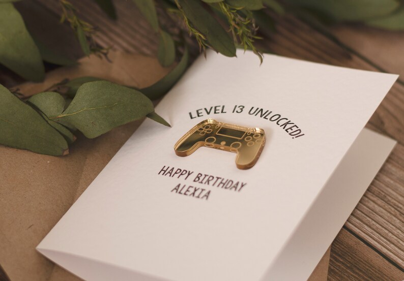 May include: A white card with the text "Level 13 Unlocked!" and a gold-colored video game controller. The card also says "Happy Birthday Alexia".