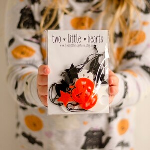 May include: A clear plastic bag containing a selection of Halloween decorations, featuring ghosts, bats, pumpkins and stars. The decorations are made from white and orange felt and are strung together with black twine. The bag is labelled "two little hearts" and includes the website address "www.two littleheartsuk.etsy.com".
