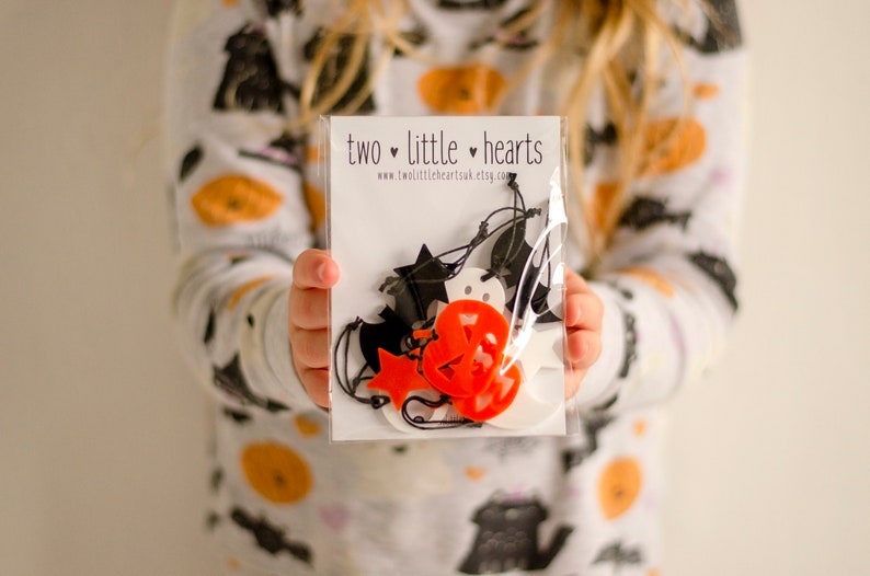 May include: A clear plastic bag containing a selection of Halloween decorations, featuring ghosts, bats, pumpkins and stars. The decorations are made from white and orange felt and are strung together with black twine. The bag is labelled "two little hearts" and includes the website address "www.two littleheartsuk.etsy.com".