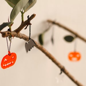 May include: A branch with three Halloween decorations hanging from it. The decorations are a black bat, an orange pumpkin, and another orange pumpkin. The branch is brown and has green leaves.