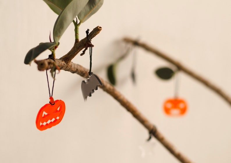 May include: A branch with three Halloween decorations hanging from it. The decorations are a black bat, an orange pumpkin, and another orange pumpkin. The branch is brown and has green leaves.