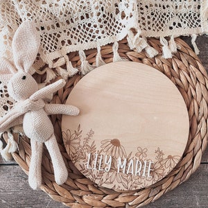 May include: A round wooden sign with a floral design and the name "Lily Marie" engraved in white lettering.