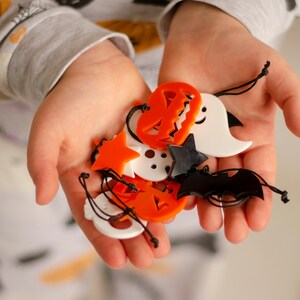 May include: A child's hand holding a variety of Halloween-themed charms, including pumpkins, ghosts, bats, and stars. The charms are made of orange, white, and black plastic.
