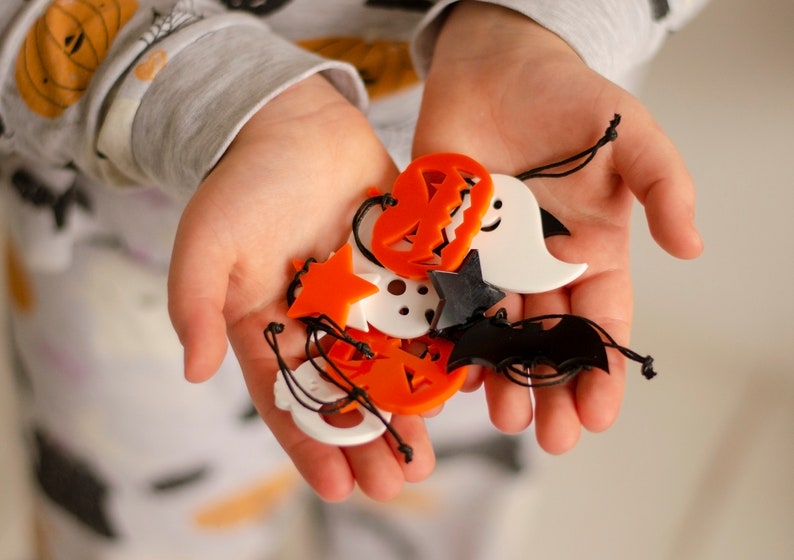 May include: A child's hand holding a variety of Halloween-themed charms, including pumpkins, ghosts, bats, and stars. The charms are made of orange, white, and black plastic.