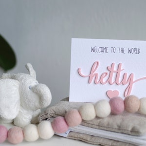 May include: A white card with the text "Welcome to the world Hetty" in pink lettering with a pink heart below the name. The card is sitting on a stack of beige fabric with a white and pink garland of felt balls.