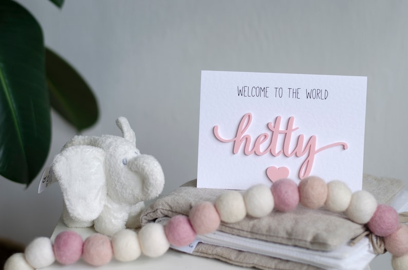 May include: A white card with the text "Welcome to the world Hetty" in pink lettering with a pink heart below the name. The card is sitting on a stack of beige fabric with a white and pink garland of felt balls.