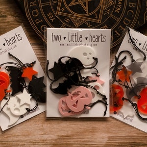 May include: Three packs of Halloween decorations. Each pack contains a variety of cut-out shapes, including pumpkins, bats, ghosts, and stars. The shapes are made of different colours, including orange, black, white, and pink. The packs are labelled "two little hearts" and include the website address www.twolittleheartsuk.etsy.com.