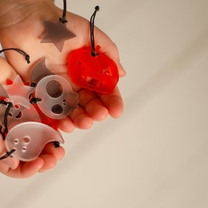 May include: A child's hand holds a variety of Halloween-themed charms, including a ghost, a skull, a bat, a pumpkin, and a star. The charms are made of translucent plastic and have black string attached to them.