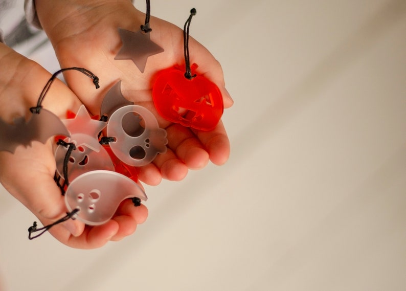 May include: A child's hand holds a variety of Halloween-themed charms, including a ghost, a skull, a bat, a pumpkin, and a star. The charms are made of translucent plastic and have black string attached to them.