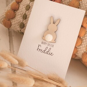 K&ouml;nnte beinhalten: Eine wei&szlig;e Karte mit einem ausgeschnittenen Holzhasen und einem wei&szlig;en Pompon-Schwanz. Die Karte sagt "Frohe Ostern Freddie".