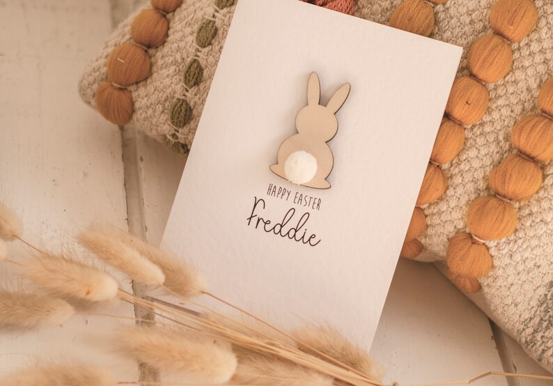 K&ouml;nnte beinhalten: Eine wei&szlig;e Karte mit einem ausgeschnittenen Holzhasen und einem wei&szlig;en Pompon-Schwanz. Die Karte sagt "Frohe Ostern Freddie".