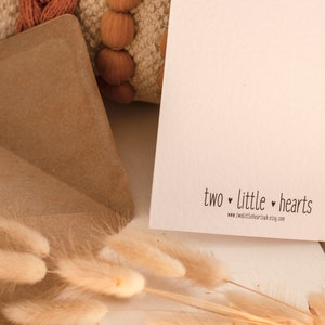 K&ouml;nnte beinhalten: Eine wei&szlig;e Karte mit dem Text "two little hearts" und der Website-Adresse "www.twolittlehearts.etsy.com". Die Karte liegt auf einer Holzoberfl&auml;che mit einem braunen Umschlag und etwas trockenem Gras.