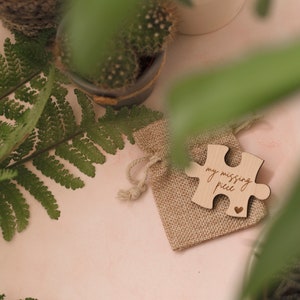 May include: A small wooden puzzle piece in a burlap bag. The puzzle piece is engraved with the words "my missing piece" and a small heart.