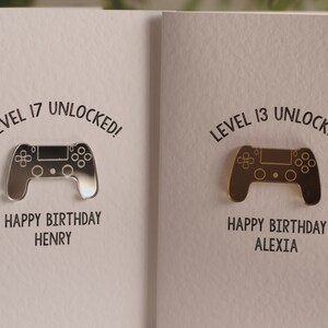 May include: Two birthday cards with a silver and gold video game controller design. The text on the cards reads "Level 17 Unlocked! Happy Birthday Henry" and "Level 13 Unlocked! Happy Birthday Alexia".