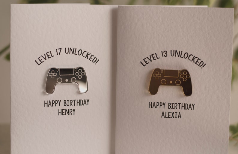May include: Two birthday cards with a silver and gold video game controller design. The text on the cards reads "Level 17 Unlocked! Happy Birthday Henry" and "Level 13 Unlocked! Happy Birthday Alexia".