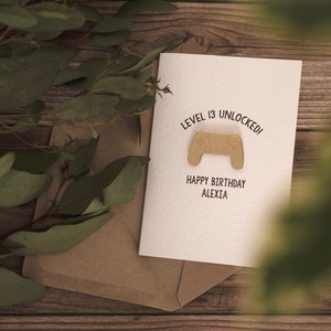 May include: A white card with the text "Level 13 Unlocked!" and a brown video game controller graphic. Below the graphic is the text "Happy Birthday Alexia".