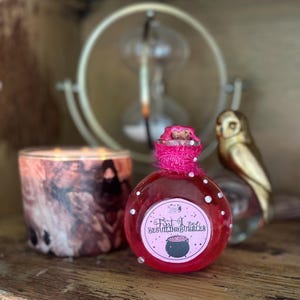 May include: A red potion bottle with a pink, textured top and a label that reads "First Love Beguiling Bubbles." The bottle is decorated with small, clear gems. A marbled candle and a gold owl figurine are also visible.