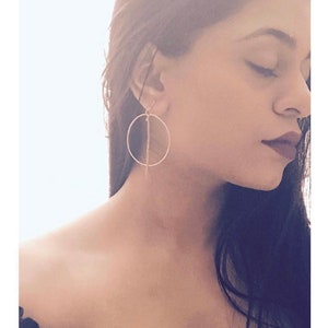 Puede incluir: Una mujer lleva un par de pendientes de aro de oro con una fina línea vertical en el centro de cada aro.