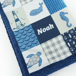 Può includere: Una copertina per neonati blu e bianca a tema nautico. La copertina presenta una balena, un cavalluccio marino, un faro e tartarughe. La copertina è personalizzata con il nome "Noah".