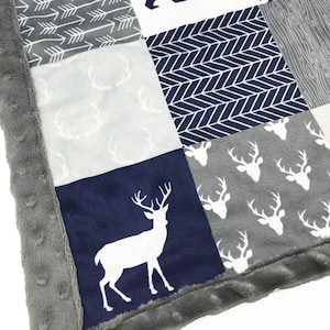 May include: A baby blanket with a patchwork design featuring a navy blue background with white deer, arrows, and a bear silhouette. The blanket has a grey border with a soft, plush texture.