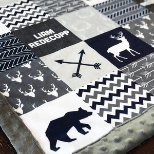 May include: A grey and navy blue baby blanket with a patchwork design. The blanket features a variety of patterns, including deer, arrows, and a bear. The blanket also has a personalised name tag that reads "Liam Redekopp".