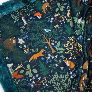 May include: A teal blue fleece blanket with a dark blue background featuring a woodland pattern. The pattern includes foxes, deer, bears, birds, mushrooms, and flowers.