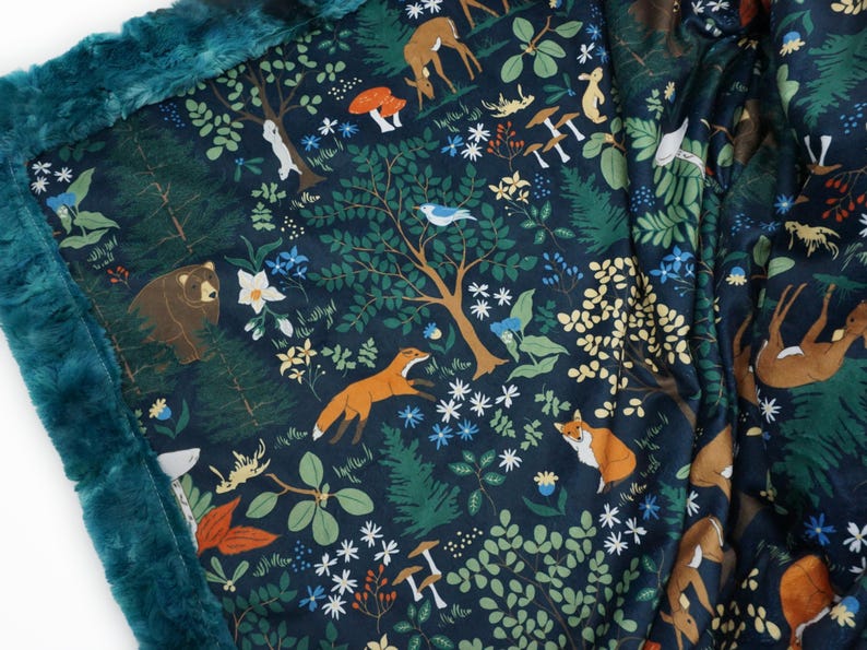 May include: A teal blue fleece blanket with a dark blue background featuring a woodland pattern. The pattern includes foxes, deer, bears, birds, mushrooms, and flowers.